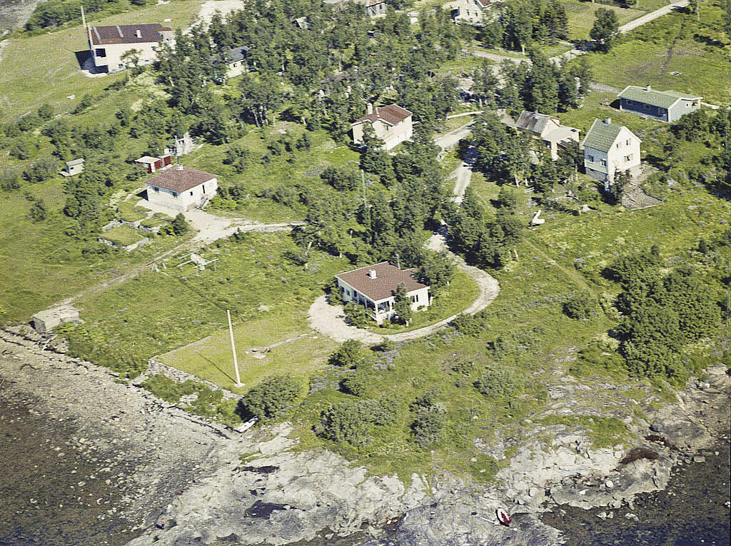 Sydspissen 1960. Sydspissen leir lå der hvor flaggstangen står. Muren ligger i overgangen mellom gress og strand. Foto: Fjellanger Widerøe, Perspektivmuseet